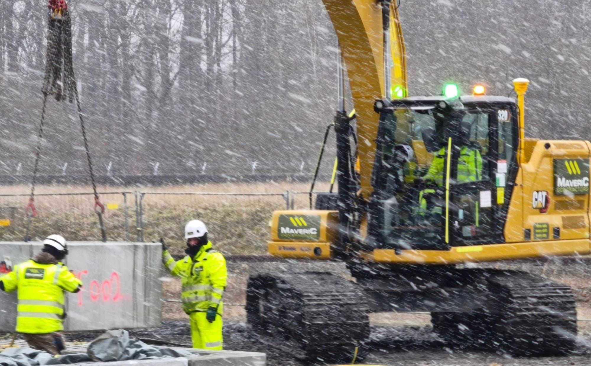 A Maveric Caterpillar excavator lifting a precast concrete unit by chains on a snowy civils site, banked by two operatives.
